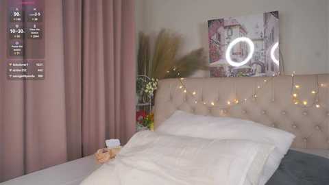 Video of a modern bedroom with beige tufted headboard, white bedding, pink curtains, a wall-mounted circular mirror, and fairy lights.