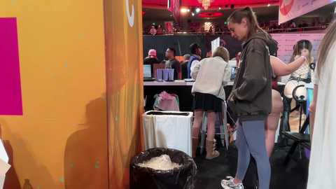 Video of a diverse group of women, some seated, some standing, at a busy event booth with yellow walls, a white trash can, and a pink banner.