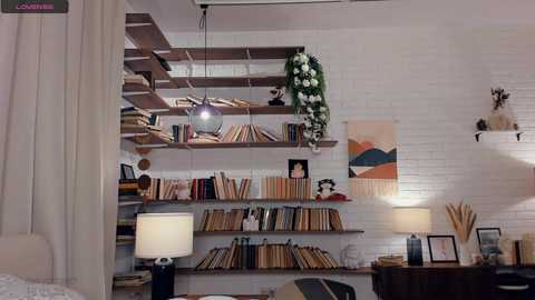 A video of a cozy, book-filled room with white brick walls, wooden shelves, hanging plants, and a modern white lamp.