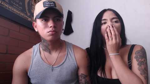 Media: Video of a young woman with long black hair, medium breasts, and tattoos covering her arms, covering her face with her hands, standing next to a man with tattoos and a beige baseball cap, wearing a grey tank top.