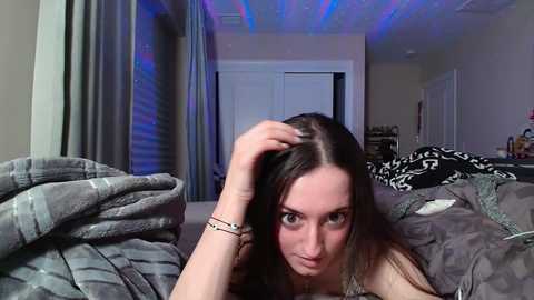 Media: Video of a young Caucasian woman with long brown hair, wearing a black top, lying on a bed with gray blankets, in a dimly lit bedroom with LED lights.