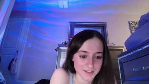 Media: Video of a young Caucasian woman with straight brown hair, wearing a gray tank top, smiling slightly. Room with blue and purple lighting, beige walls, dresser, and closed door in the background.