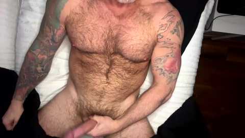 Media: Video of a muscular, hairy, light-skinned man with tattoos, lying nude on a bed, holding his erect penis. Background includes a wooden floor and a partially visible TV.