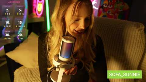 Media: Video of a young blonde girl with long hair, smiling while holding a blue light-up microphone, in a dimly lit room with colorful neon lights and a sofa.