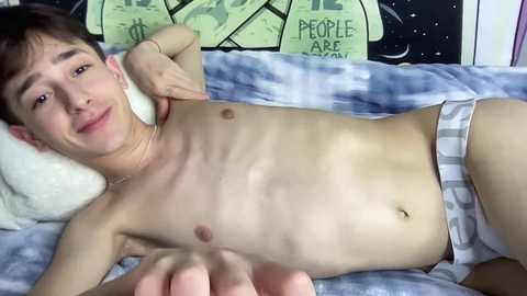 Media: Video of a young, fair-skinned, slim, shirtless boy lying on a bed with gray plaid boxers, smiling, with a white pillow and green \"People Are\" poster in the background.