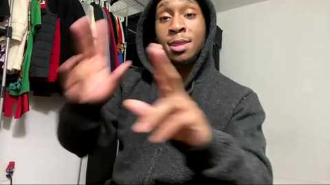 Media: Video of a young Black man in a dark hoodie, hands in a \"peace\" sign, standing in a cluttered closet with hanging clothes.