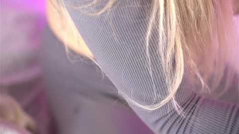 Media: Video of a young woman with long, blonde hair, wearing a grey ribbed sweater, with blurred background featuring pastel colors and soft lighting.