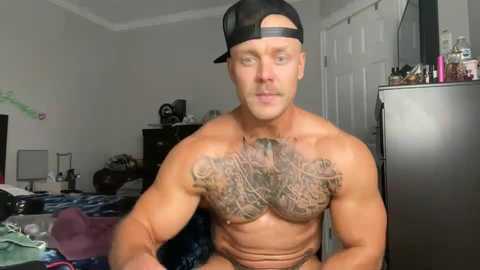 Media: Video of a shirtless, muscular, bald man with a black cap, wearing a large, intricate tattoo across his chest. He sits in a messy bedroom with a fridge, dresser, and bed visible.