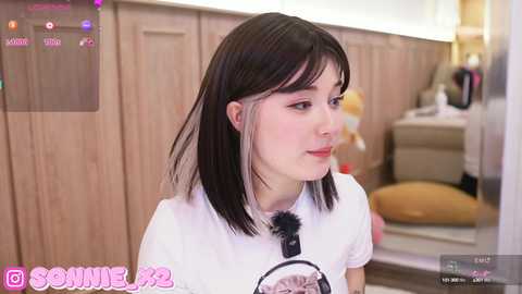 Media: A video of a young woman with straight, shoulder-length dark hair, wearing a white t-shirt, standing in a modern, beige-toned kitchen.