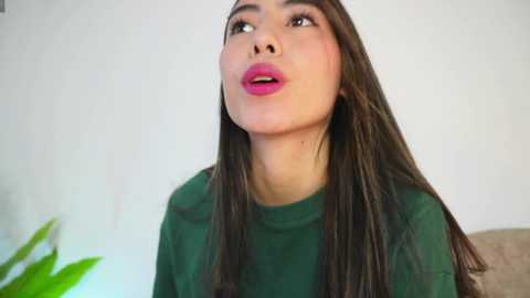 Media: Video of a young woman with long, straight brown hair, wearing bright pink lipstick and a green top, gazing upward in a casual indoor setting.