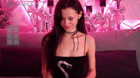 Media: Video of a slender, fair-skinned woman with long black hair, wearing a black top with a snake graphic and a black choker, standing in front of a pink-lit, decorative Christmas tree background.