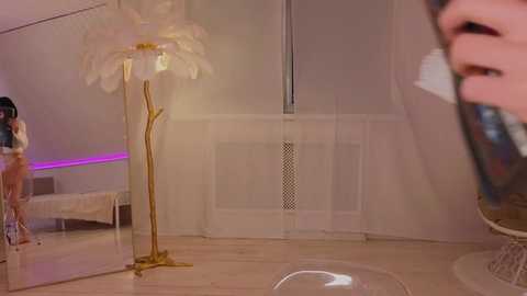 A video of a modern, minimalist bedroom featuring a tall, golden feather lamp, a white bed with a purple accent strip, and a blurry reflection of a person with a tattooed arm.