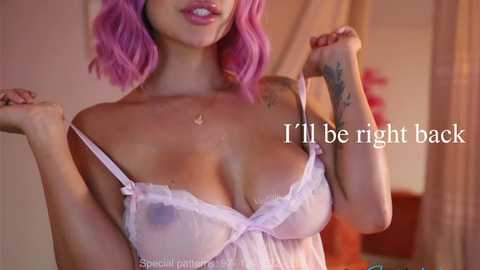 Video of a light-skinned woman with vibrant pink hair, wearing a sheer, lacy, lavender lingerie top, revealing her large breasts. Text overlay reads, \"I'll be right back.\