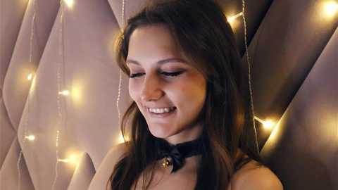 Media: Video of a smiling young woman with long brown hair, wearing a black bow necklace, against a backdrop of a quilted, padded wall adorned with golden fairy lights.