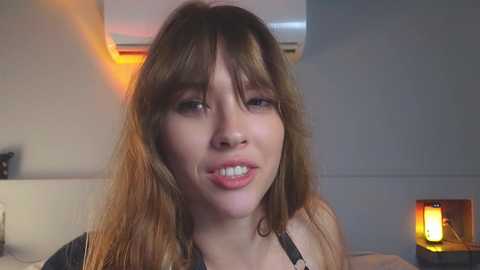 Media: Video of a young Caucasian woman with light skin, long brown hair, and a playful expression. She wears a black top. Background shows a beige wall, a lamp, and a white couch.