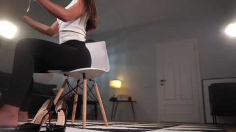 Media: Video of a woman in a white blouse and black pants seated at a modern white chair with wooden legs, facing a white door in a minimalist, dimly lit room with a black-and-white checkered rug.