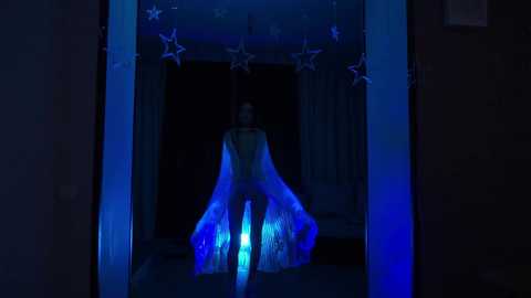 Video of a dark room with a lit-up, glowing figure dressed in a flowing, ethereal gown, surrounded by star-shaped lights. The scene is bathed in blue hues, creating a mystical, dreamlike atmosphere.