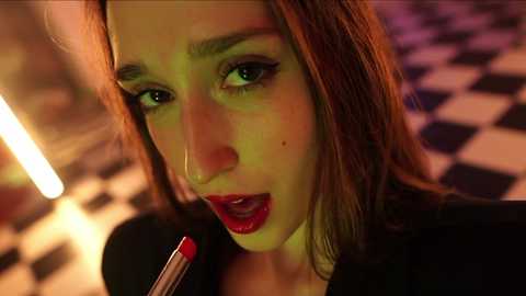 A close-up video of a young woman with light brown hair and red lipstick, holding a red lipstick, in a dimly lit, checkered room, evoking a retro diner vibe.