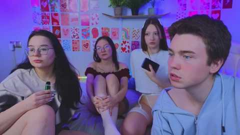 Media: Video of four young people in a colorful room with a collage of hearts on the wall. Two girls wear glasses, one in a white dress, another in a red top. A boy in a blue jacket sits, looking serious.