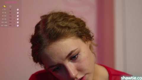 Media: Video of a young woman with light skin, curly brown hair, and subtle makeup, wearing a red top, looking downward. Background features a blurred pink wall and a calendar interface with the date \"1/1\" visible.