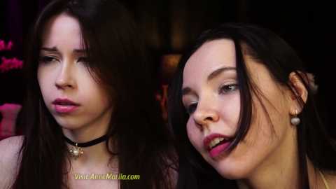 Media: Video of two young women with long dark hair, one wearing a black choker, the other pearl earrings, both with fair skin, appearing emotional, set against a dimly lit, blurred background.