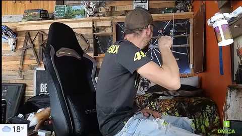 A video of a muscular, fair-skinned man with glasses and a brown cap, wearing a black \"DNR\" t-shirt, seated on a black gaming chair, playing a video game in a rustic, cluttered room.