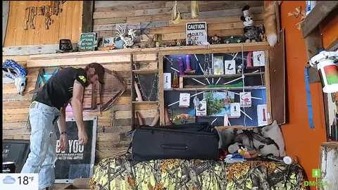 Media: Video of a woman with long hair in a black shirt and jeans adjusting a camouflage couch in a rustic, wooden-paneled room with deer antlers and animal-themed decor.