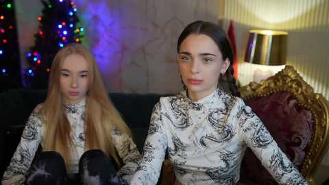 Media: Video of two young women with long, straight hair, dressed in white, patterned tops, sitting on a dark couch in a cozy room with a lit Christmas tree, festive lights, and a gold armchair.