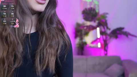 Media: A video of a young woman with long, wavy brown hair, wearing a black top, smiling. The background features a blurred living room with greenery, purple lighting, and a gray couch.