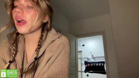 Media: Video of a young Caucasian woman with blonde hair in braids, wearing a beige ribbed sweatshirt, caught in a candid moment. Background includes a white door with a black \"STREET\" towel hanging.