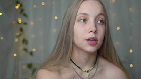 Video of a young, fair-skinned, blonde-haired woman with blue eyes, wearing a black choker necklace, against a softly lit, blurred background with greenery and fairy lights.