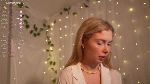 Media: Video of a young blonde woman with fair skin, wearing a white blouse, and a beaded necklace, looking down. Background features soft, glowing fairy lights and green ivy.