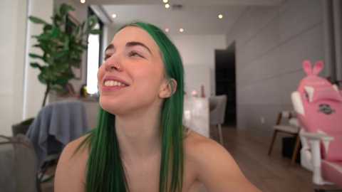 Media: Video of a smiling young woman with long, vibrant green hair, standing in a modern, brightly lit room with minimalist decor.