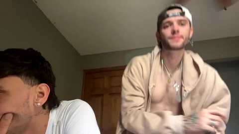 Media: Video of two young men in a casual indoor setting; one with short dark hair, white T-shirt, and laughing, the other with a backward cap, beige jacket, and necklace.