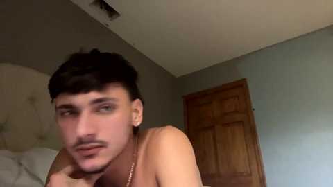 Media: A video of a shirtless young man with short dark hair, light skin, and a trimmed mustache, lying on a bed, looking contemplative, in a simple, dimly lit bedroom with a wooden door and beige walls.