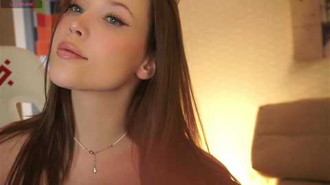 Video of a young, fair-skinned woman with long brown hair, wearing a silver necklace, standing in a warmly lit room with beige walls, blurred background, and soft focus.