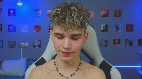 Media: Video of a shirtless young man with curly, dyed hair, wearing a black beaded necklace, sitting in a gaming chair in a dimly lit room with blue lighting and posters on the wall.