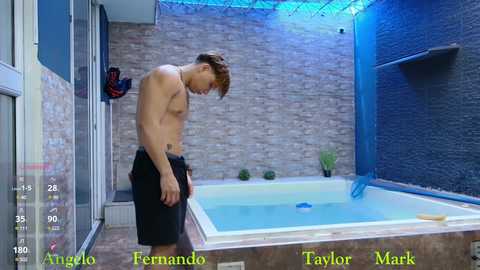 Video of shirtless, muscular young man with light skin, wearing black shorts, standing by a small blue-tiled indoor pool with a brick wall background.