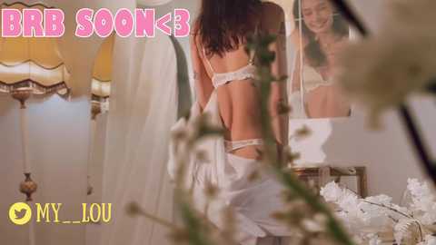 Media: Video of a woman with long dark hair, wearing white lace lingerie, standing in front of a mirror in a softly lit, vintage-style bedroom. Text reads \"BRA SOON\" and \"MY LOU.\