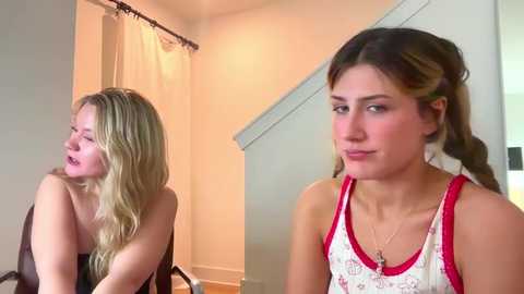 Media: Video: Two young women, one blonde with wavy hair in a black strapless top, and one brunette in a red and white floral top, sit on a white couch in a modern, beige-toned living room.