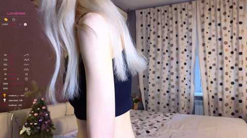 Media: Video of a slender, fair-skinned woman with long, platinum blonde hair, wearing a black crop top, standing in a bedroom with white polka-dot curtains and a bed with white sheets.