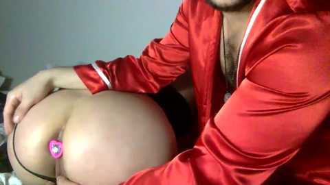 Video of a man with a light skin tone, wearing a red satin robe, holding a woman's buttocks, revealing a pink heart-shaped butt plug.