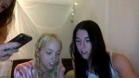 Media: Video of two young women with long hair, one blonde and one brunette, seated indoors. The blonde wears a pink shirt, while the brunette is in a white top. Both appear focused on their smartphones.