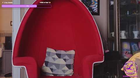 Media: A video of a modern, red, egg-shaped chair with a geometric-patterned cushion in a cozy, cluttered room with a bookshelf, kitchen, and a purple neon light.