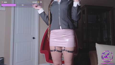 Media: Video of a woman in a tight, grey zip-up jacket and pink mini skirt, standing in a dimly lit room with a closed door, purple overlay, and a watermark.