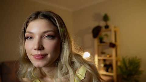 Media: Video of a young Caucasian woman with long blonde hair and fair skin, wearing a yellow top, smiling softly in a warmly lit room with a wooden bookshelf and green plants.