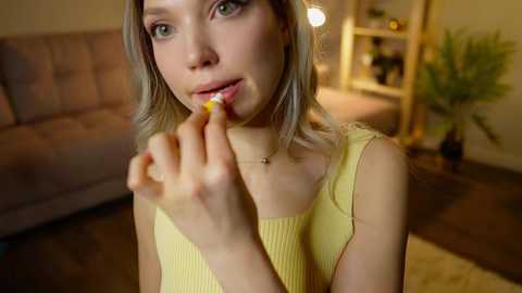 Media: Video of a young woman with light skin and shoulder-length blonde hair, wearing a yellow sleeveless top, licking a popsicle in a cozy, warmly lit living room with a beige sofa, wooden floor, and potted plants.