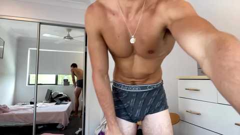 Media: Video of a shirtless, muscular man wearing Calvin Klein boxers, standing in a modern, bright bedroom with mirrored closet, dresser, and another person in the background.