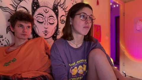 Media: Video of a young woman with glasses and a boy, both wearing casual clothes, sitting side by side in a dimly lit room with a sun tapestry and rainbow light.