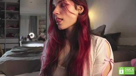 Media: Video of a young woman with long, wavy red hair, wearing a light pink blouse, in a dimly lit bedroom with a bed, shelves, and a mirror.